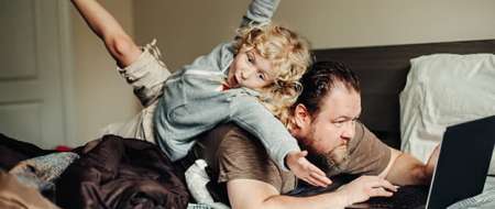 Man lying on bed using laptop while young boy plays on his back